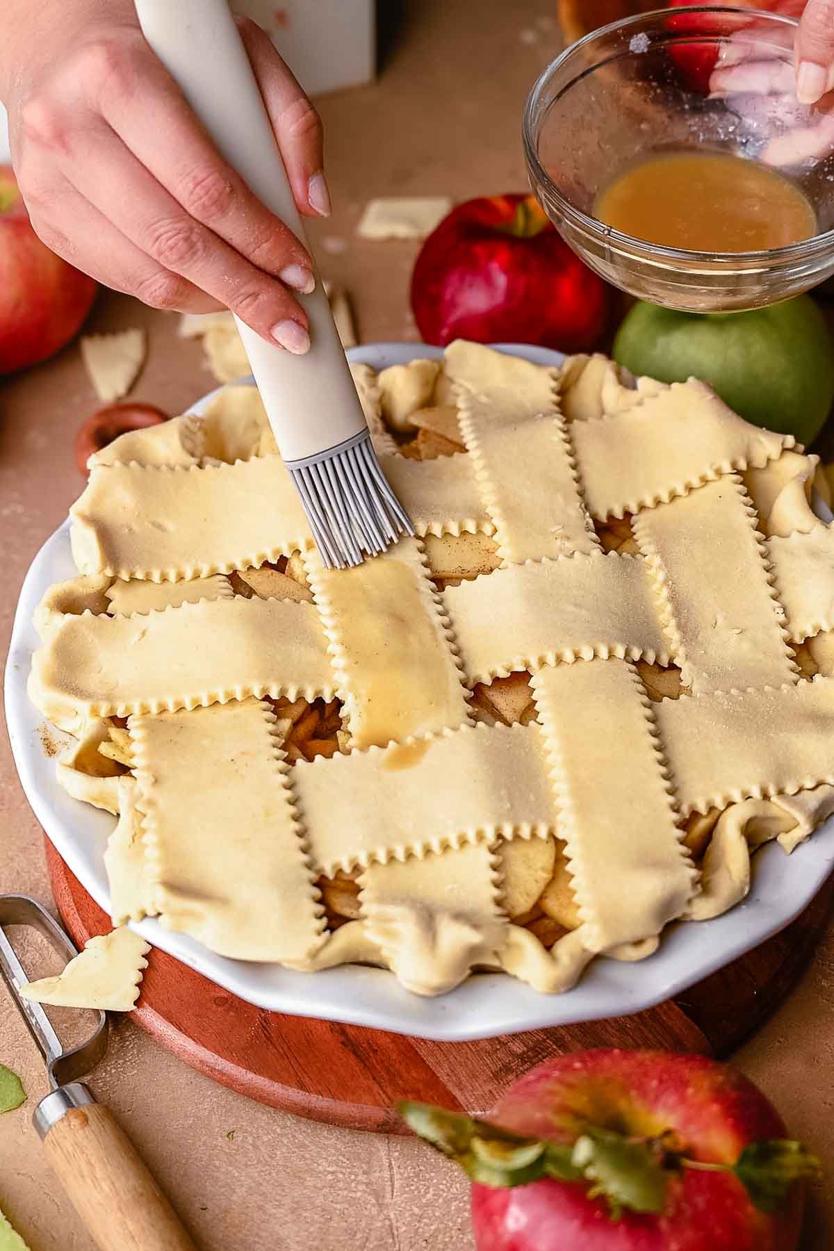 Close-up of hands brushing a lattice pie crust with vegan egg wash using a silicone pastry brush, next to a bowl of wash.