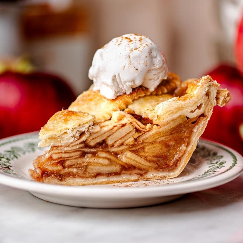 A slice of vegan apple pie with a golden flaky crust, stacked neatly with tender apple slices and topped with a scoop of vanilla non-dairy ice cream.