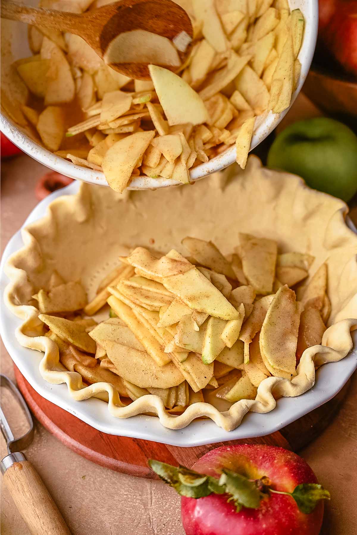 Action shot of spiced apple slices being spooned from a mixing bowl into a crimped vegan pie crust.