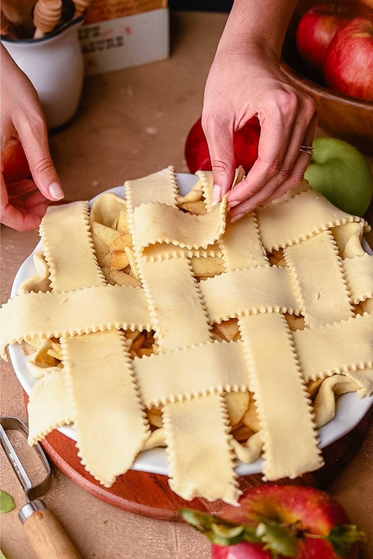 Hands folding back a strip of pie dough while weaving lattice top crust for apple pie, showing fluted-edge pastry strips.