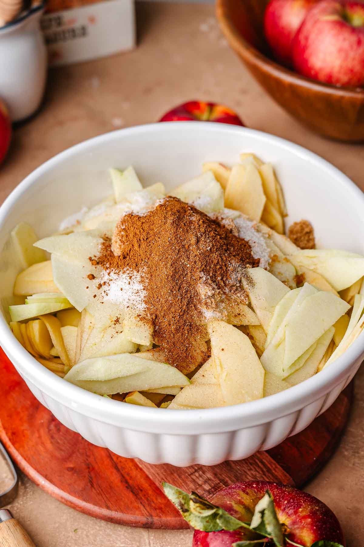 Large white mixing bowl filled with thinly sliced apples topped with brown sugar, white sugar, salt, cornstarch, and apple pie spice blend, ready to be stirred for apple pie filling.
