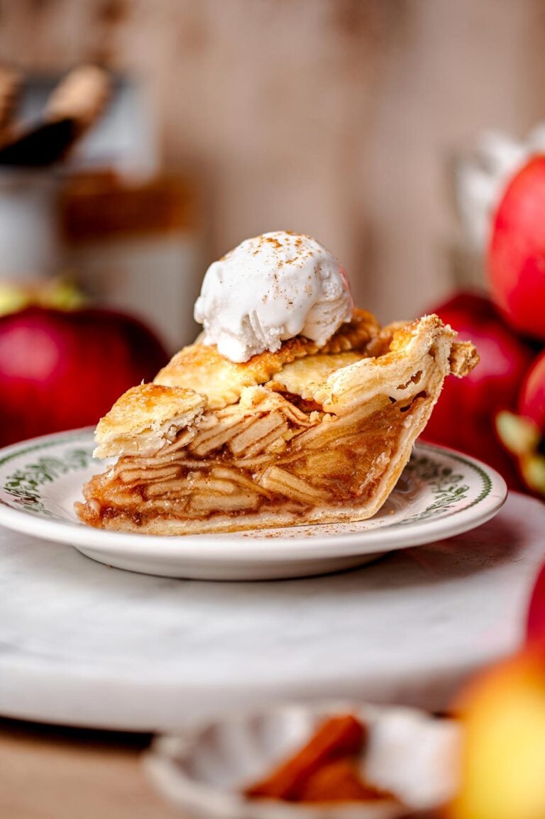 A slice of vegan apple pie with a golden flaky crust, stacked neatly with tender apple slices and topped with a scoop of vanilla non-dairy ice cream.