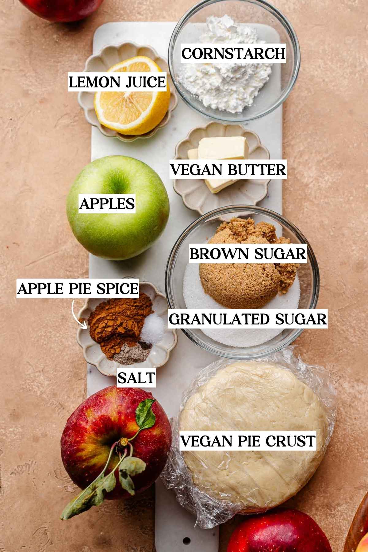 Overhead view of vegan apple pie ingredients on a marble board, including Granny Smith apple, lemon, cornstarch, vegan butter, brown sugar, white sugar, salt, apple pie spice, and wrapped vegan pie crust.