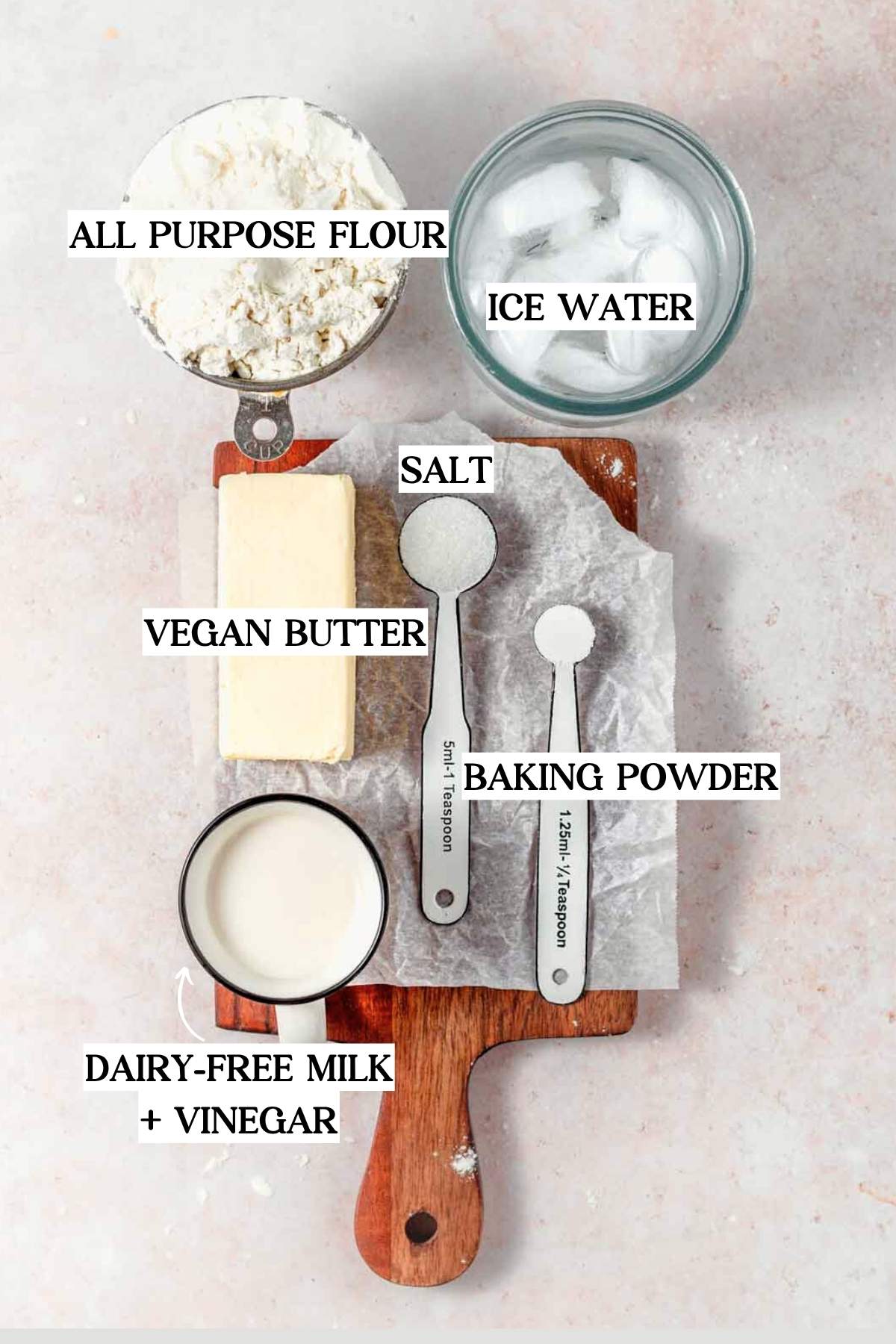 vegan pie crust ingredients on a cutting board: all-purpose flour, vegan butter, salt, baking powder, ice water, and a cup of dairy-free milk mixed with vinegar.