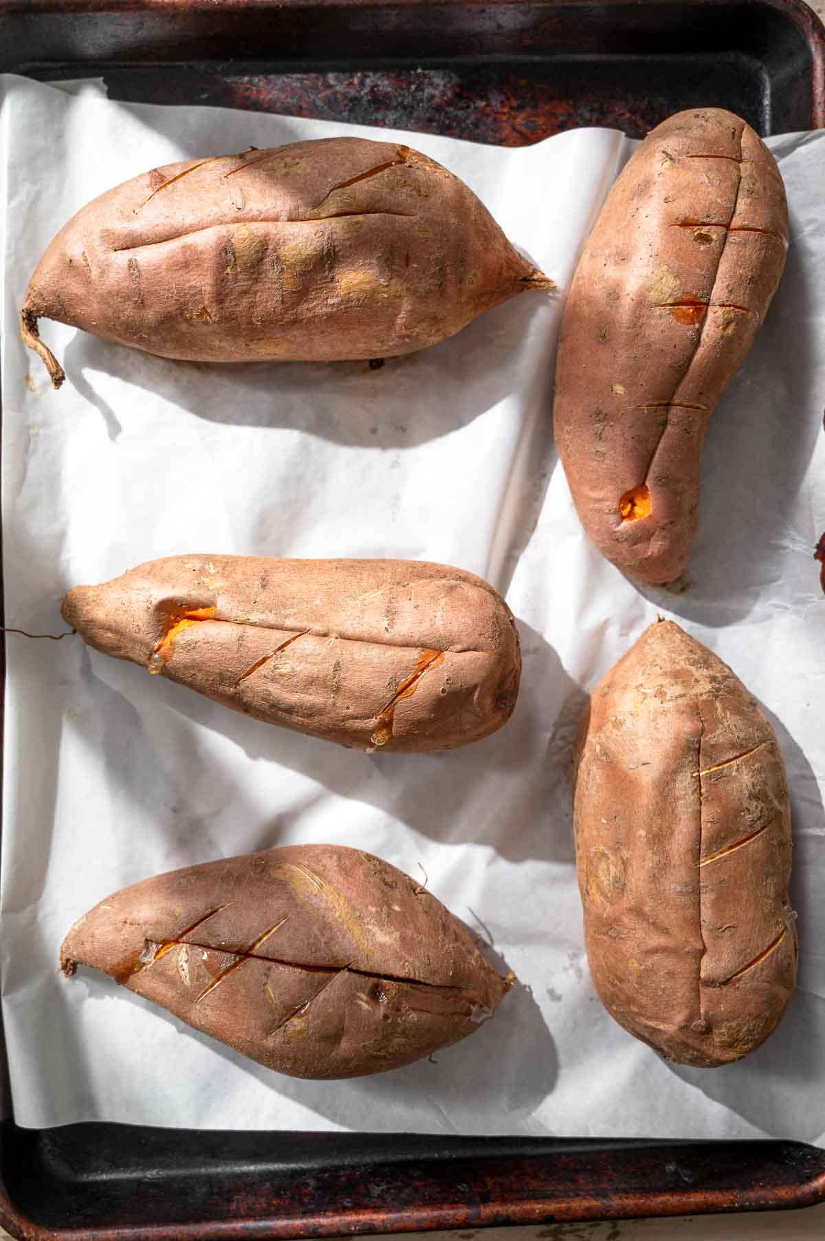 a tray with five large sweet potatoes on white parchment paper that have been baked.