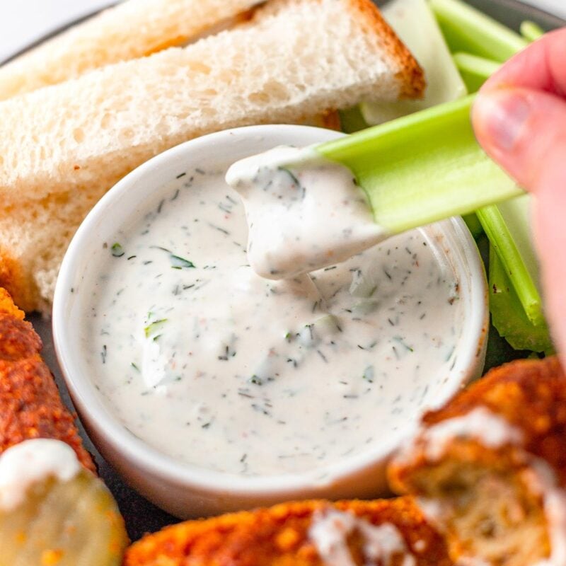 a hand dipping a celery stick in a bowl of homemade vegan ranch dressing.