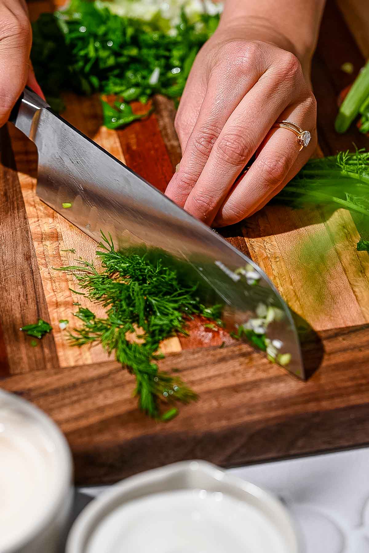 a hand finely mincing dill on a wooden cutting board.