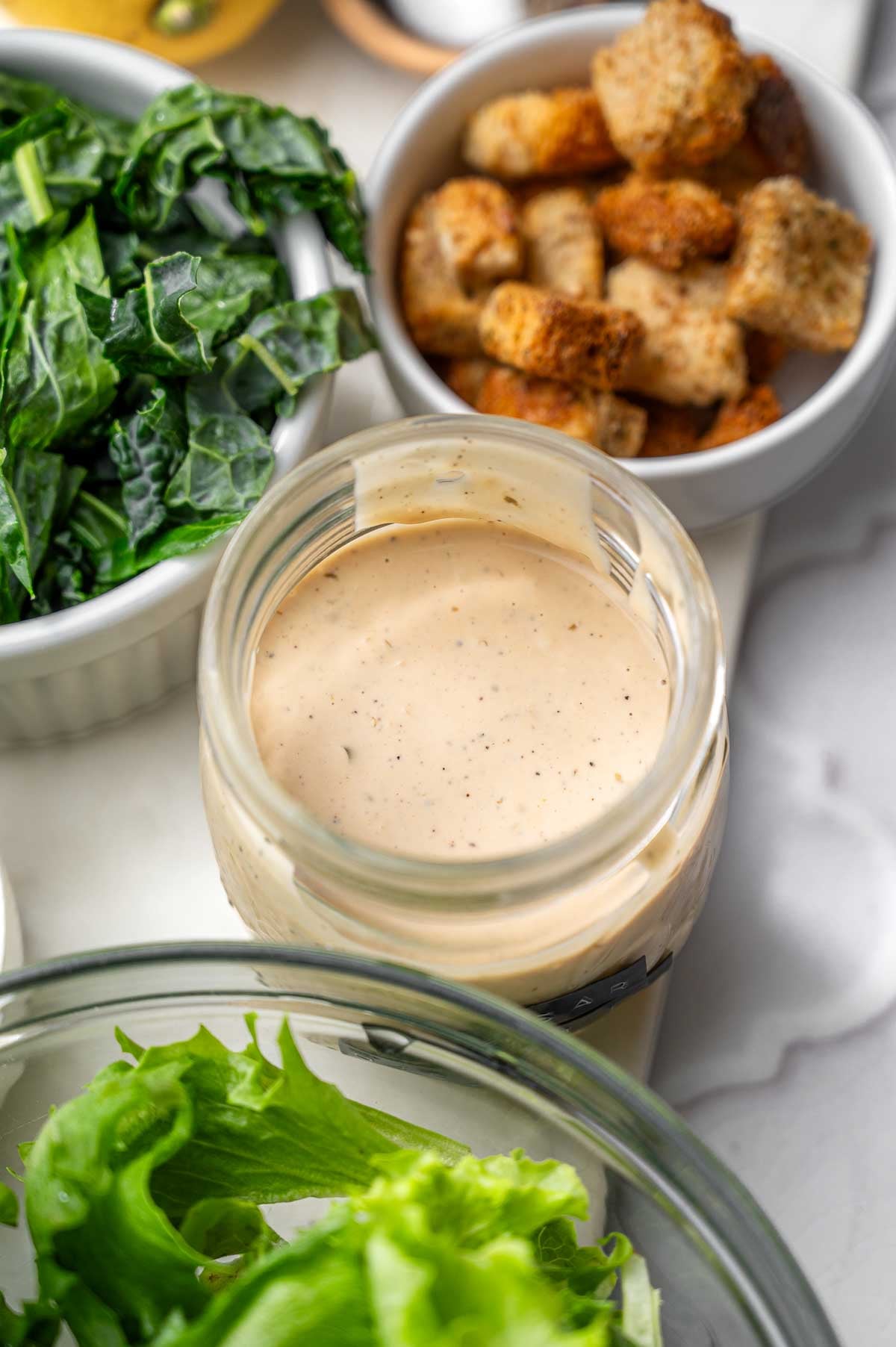 a jar of vegan caesar dressing next to lettuce and croutons.