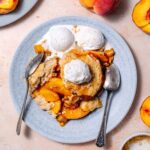 vegan peach cobbler on a blue ceramic plate with two silver spoons and three scoops of vanilla ice cream on top.