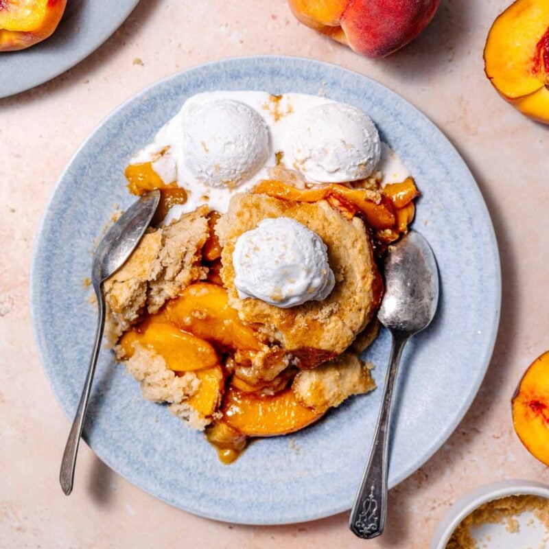 vegan peach cobbler on a blue ceramic plate with two silver spoons and three scoops of vanilla ice cream on top.
