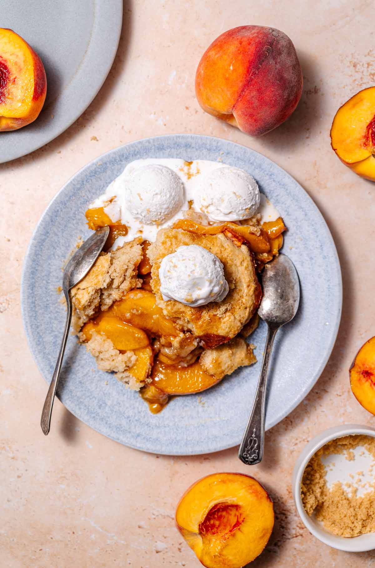 vegan peach cobbler on a blue ceramic plate with two silver spoons and three scoops of vanilla ice cream on top.