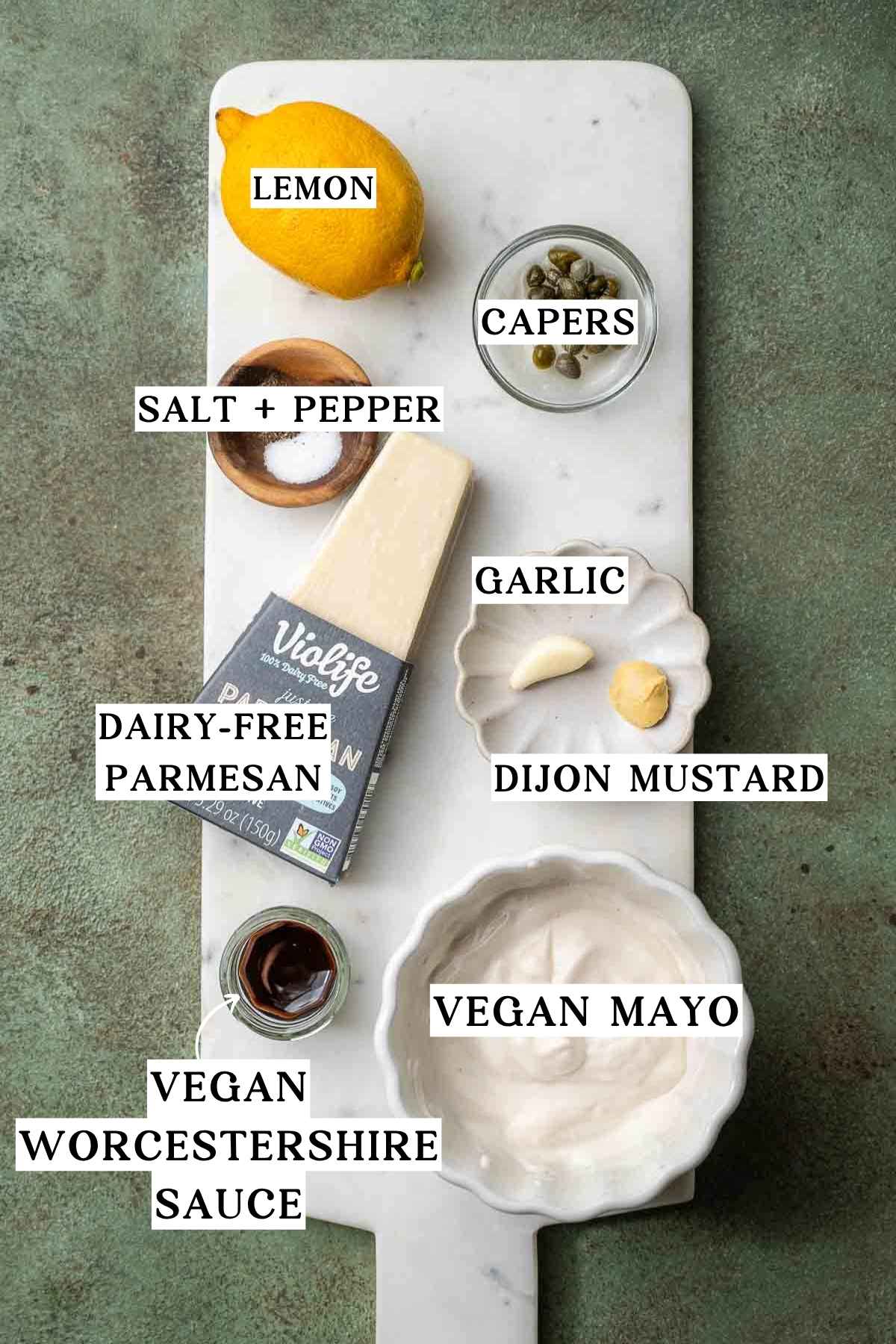 ingredients like vegan mayo, dairy-free parmesan, capers, and lemon on a marble cutting board.
