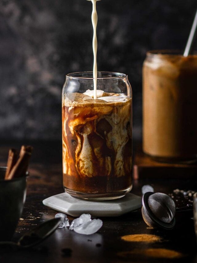 a pumpkin brown sugar shaken espresso in a serving glass with a hand pouring oatmilk into it.