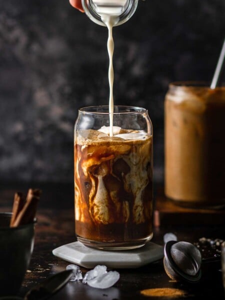 a pumpkin brown sugar shaken espresso in a serving glass with a hand pouring oatmilk into it.