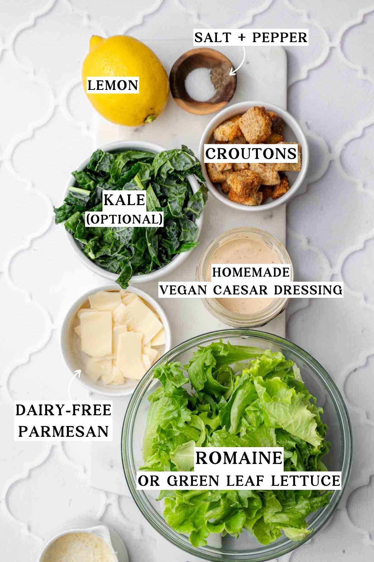 ingredients like romaine, croutons, vegan parm, lemon and caesar dressing on a marble cutting board.
