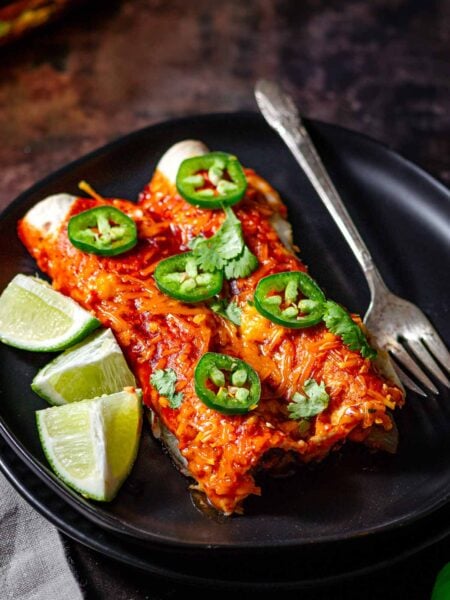 two vegan enchiladas on a black plate with lime wedges.