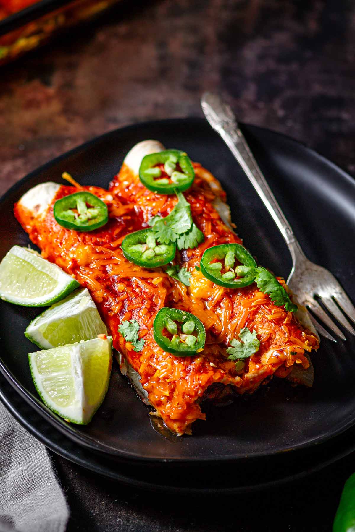 two vegan enchiladas on a black plate with lime wedges.