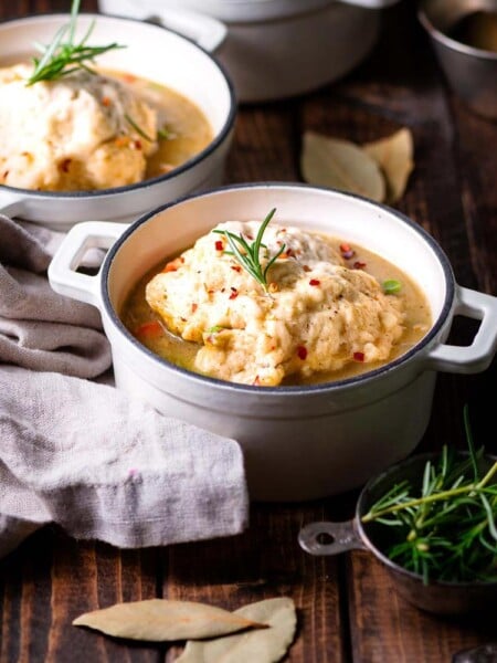 Focus shot of a white cocotte with Vegan Chicken and Dumplings on a wooden surface with another cocotte with the same dish behind