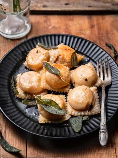 pan fried vegan butternut squash ravioli with crispy sage on a scalloped black plate.