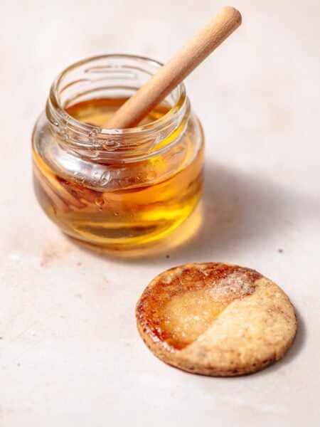 A small glass jar of golden syrup with a wooden honey dipper, next to a round piece of pastry that has been brushed with a glossy egg wash substitute and baked until golden brown.