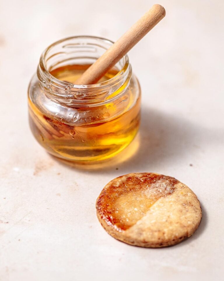 A small glass jar of golden syrup with a wooden honey dipper, next to a round piece of pastry that has been brushed with a glossy egg wash substitute and baked until golden brown.