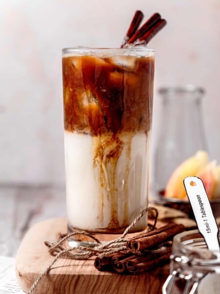 an apple crisp oatmilk macchiato in a tall glass with cinnamon sticks on top.