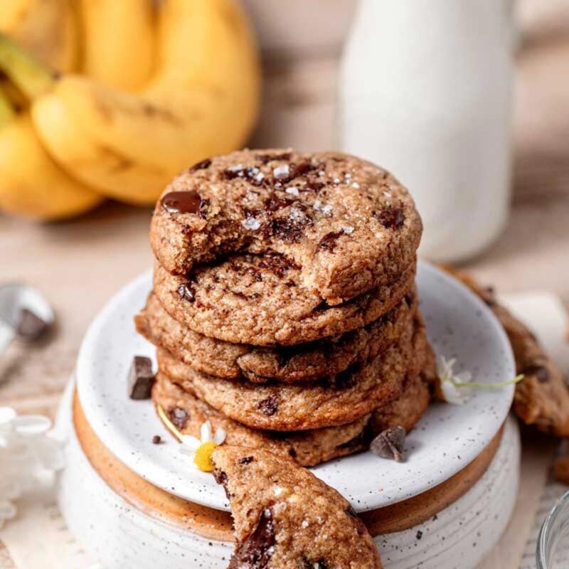 a stack of chewy vegan banana bread cookies on a white speckled ceramic plate with a jug of milk in the background.