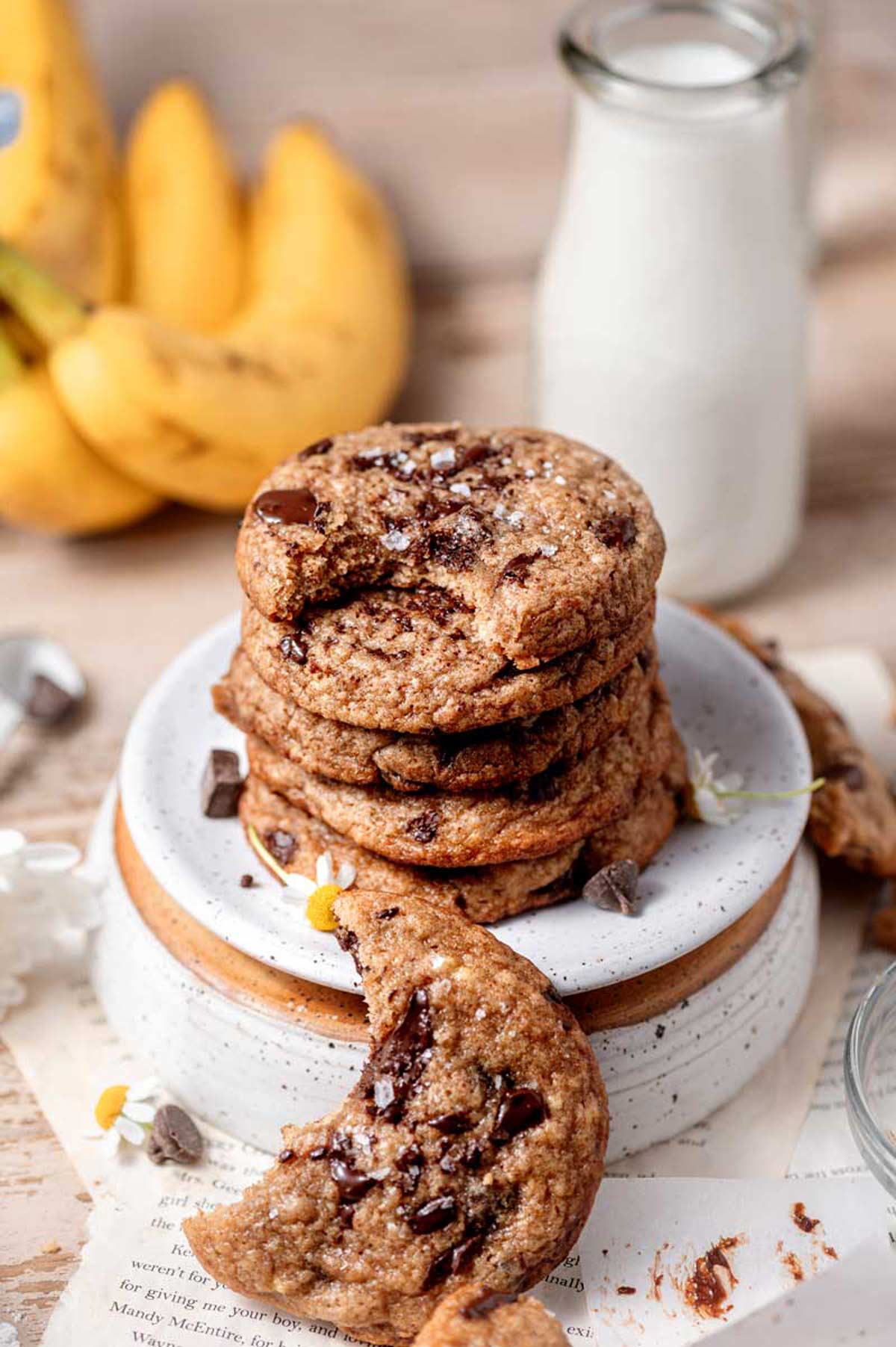 a stack of chewy vegan banana bread cookies on a white speckled ceramic plate with a jug of milk in the background.