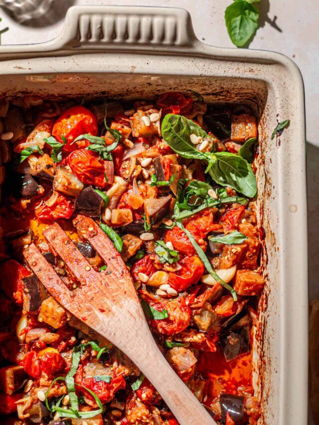 roasted eggplant and burst cherry tomatoes in a baking dish with a wooden spatula.