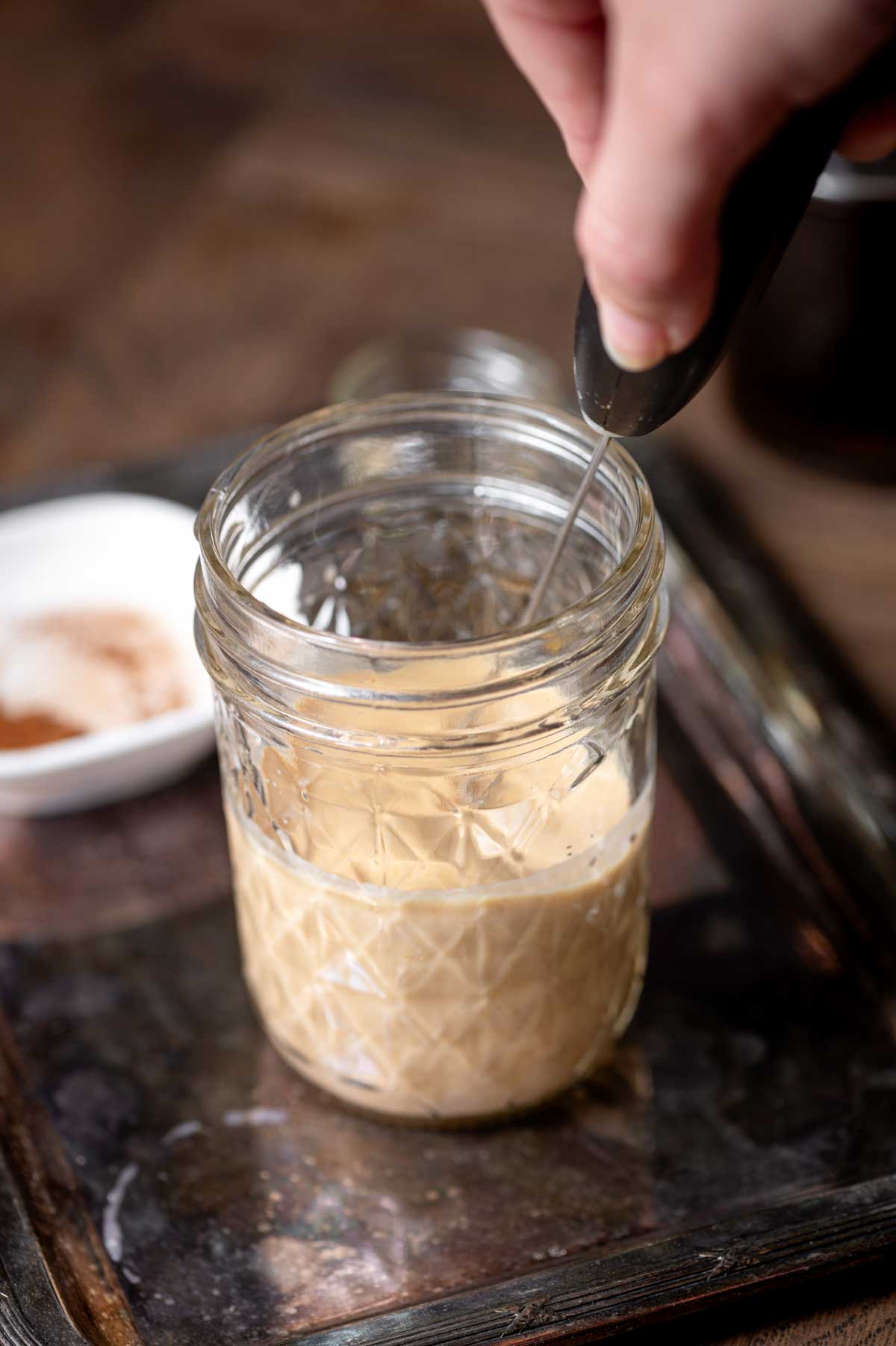 a hand using a frother to blend the pumpkin sweet cream in a small glass jar.