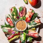 A white serving plate with colorful vegan summer rolls placed around a small bowl of peanut dipping sauce.