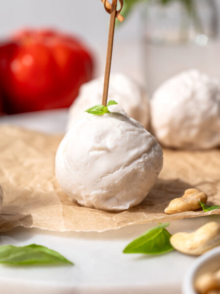 Homemade dairy-free mozzarella ball on parchment with fresh basil, cashews, tomatoes, and additional mozzarella in the background.