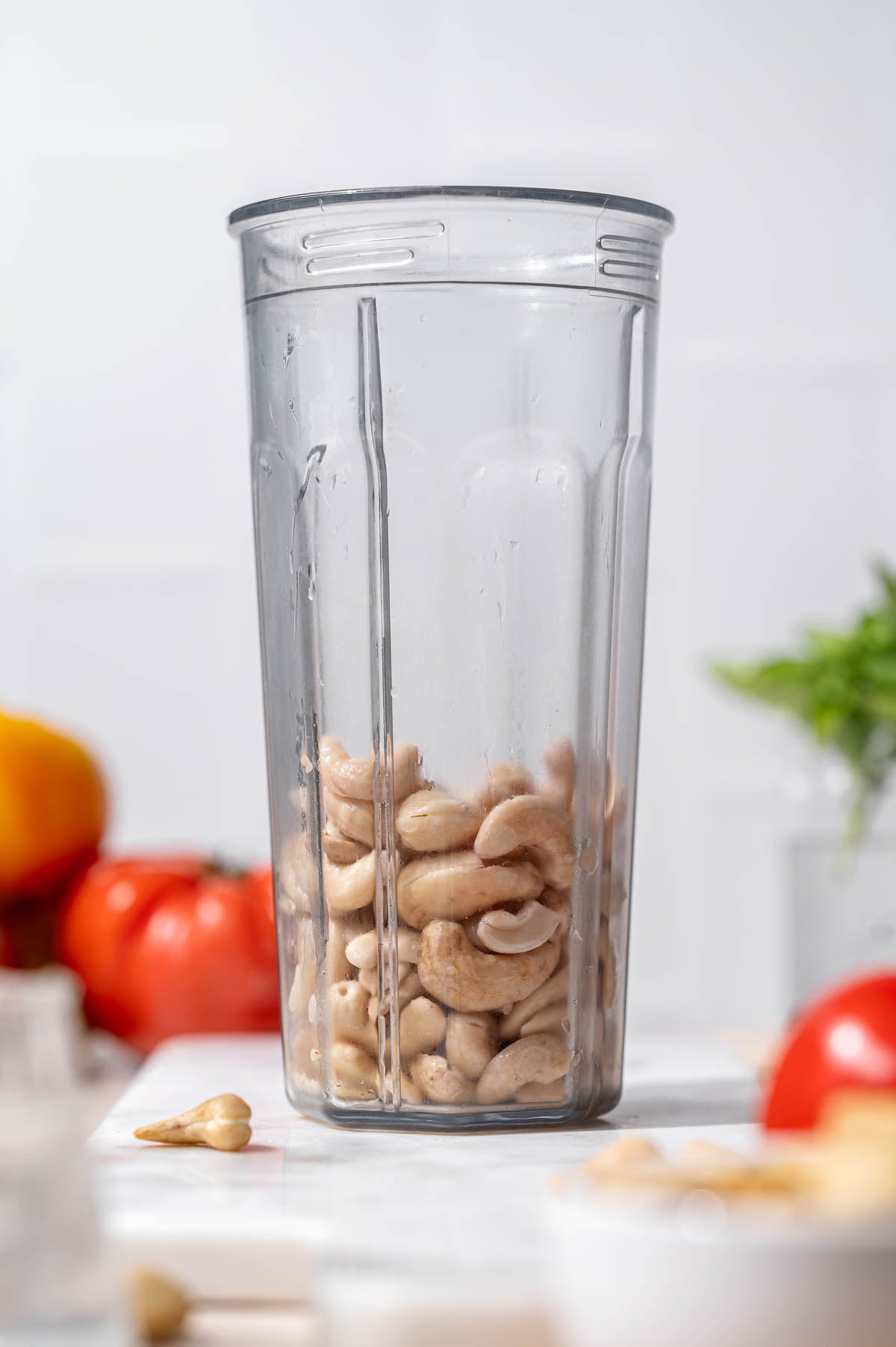 Soaked cashews in a clear blender cup with tomatoes and basil softly blurred in the background.