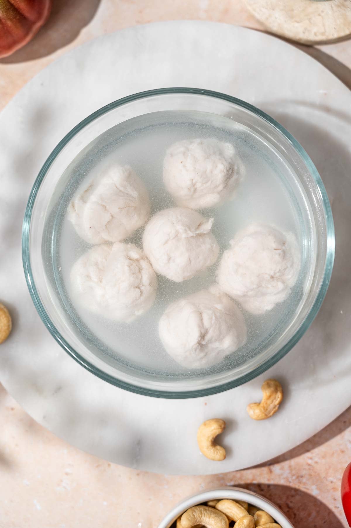 Fresh vegan mozzarella balls chilling in a glass bowl of ice water with cashews scattered nearby.