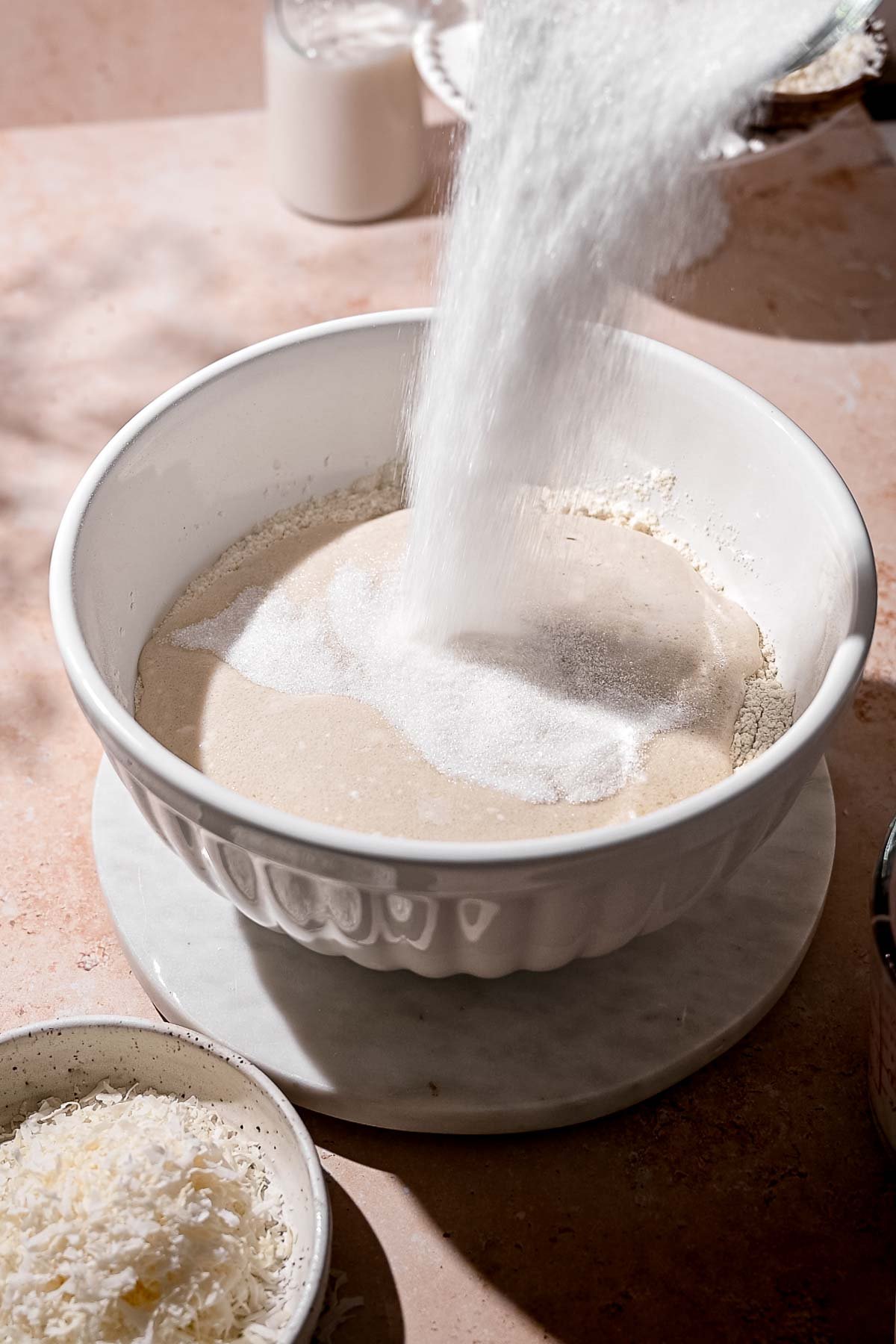 sugar being poured into a large white mixing bowl with flour and coconut milk.