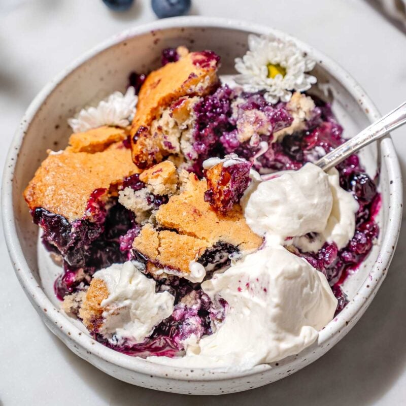 vegan blueberry cobbler with whipped cream in a white ceramic dish.