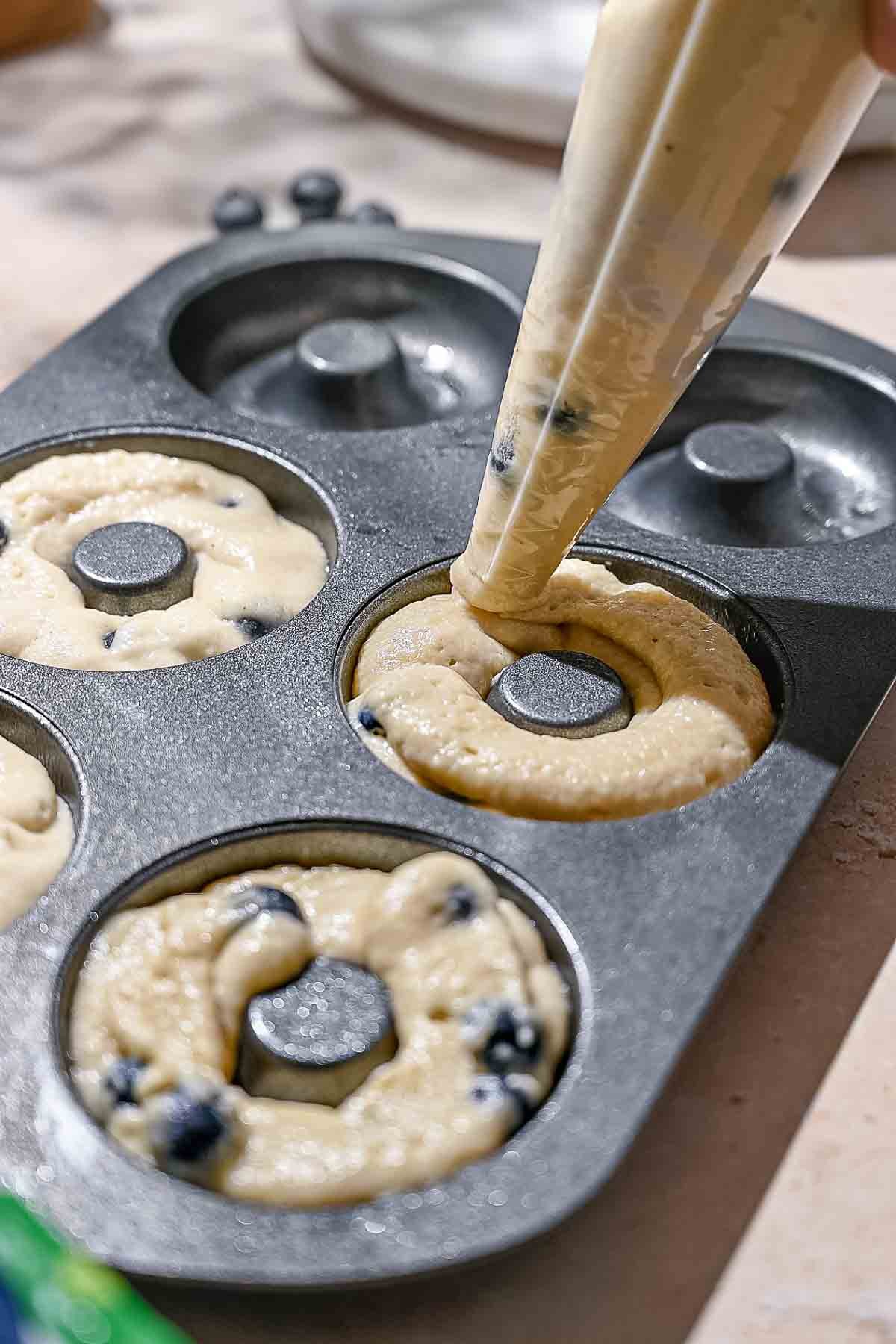a piping bag piping blueberry donut batter into a donut pan.