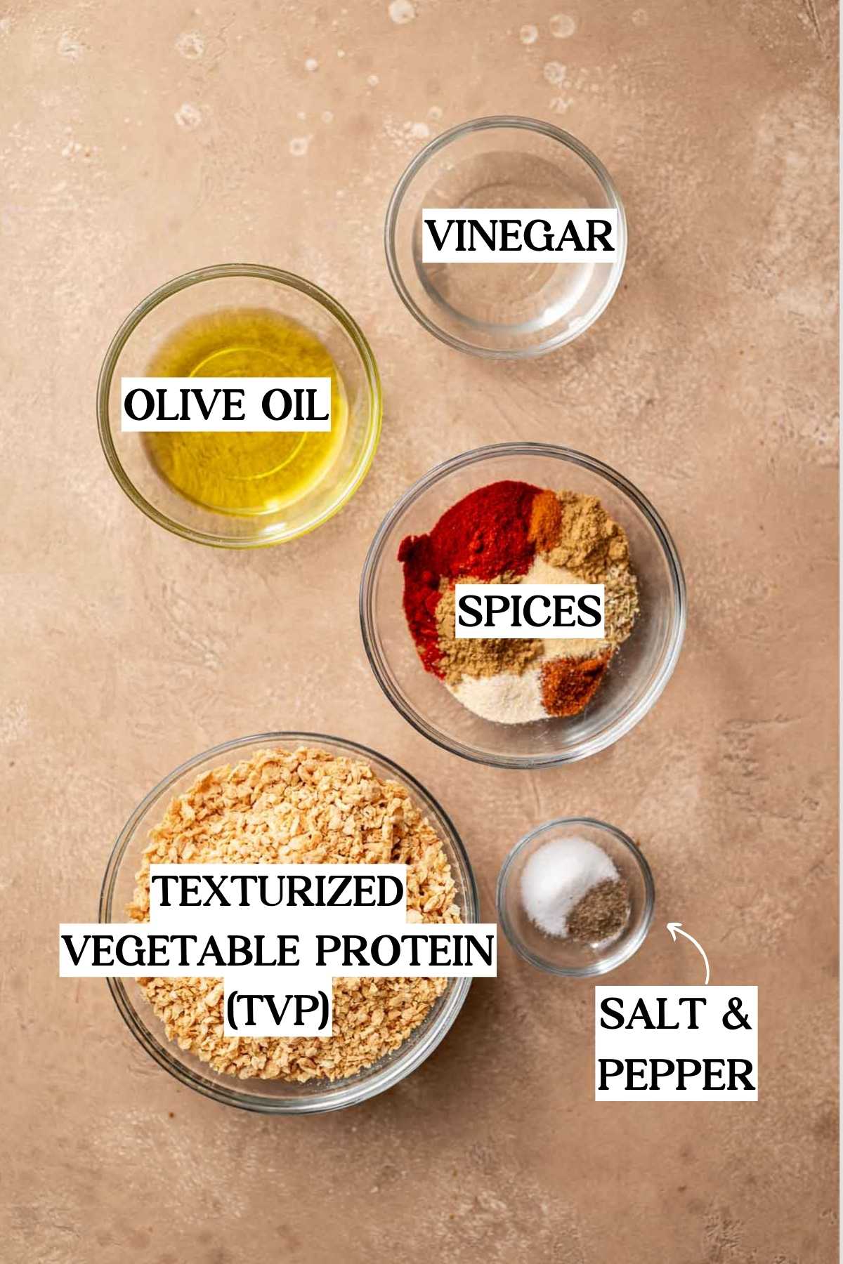 ingredients like TVP crumbles, spices, paprika and more in glass bowls on a tan surface.