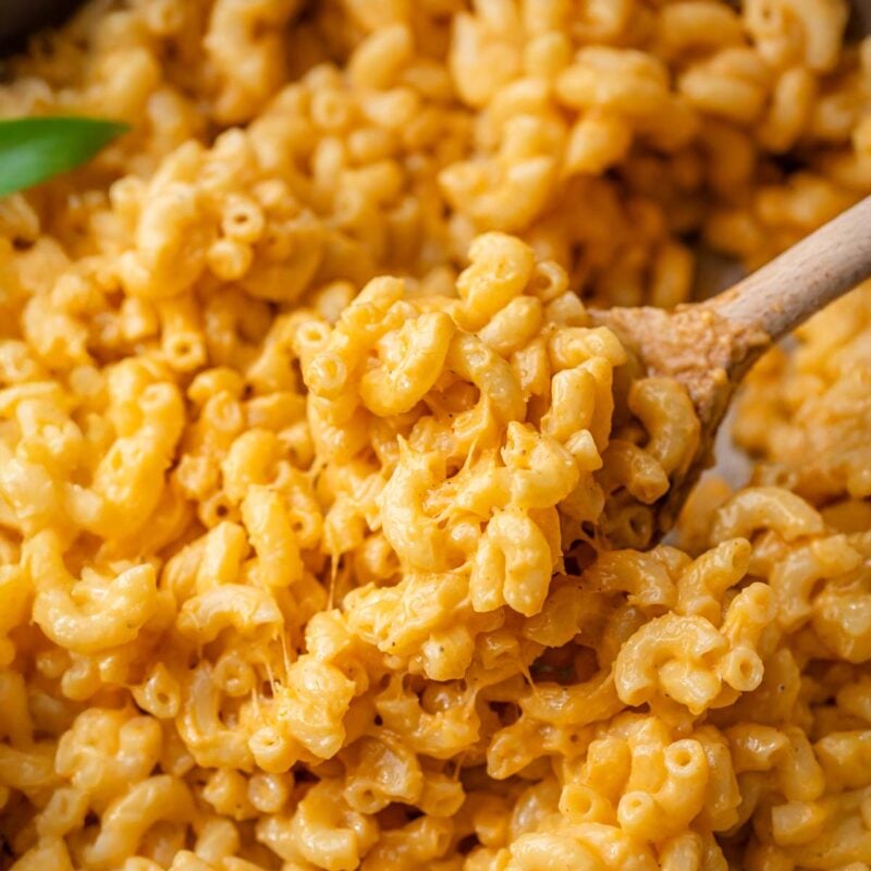 Close-up of creamy vegan mac and cheese with elbow pasta, coated in a rich, stretchy orange cheese sauce, stirred with a wooden spoon.