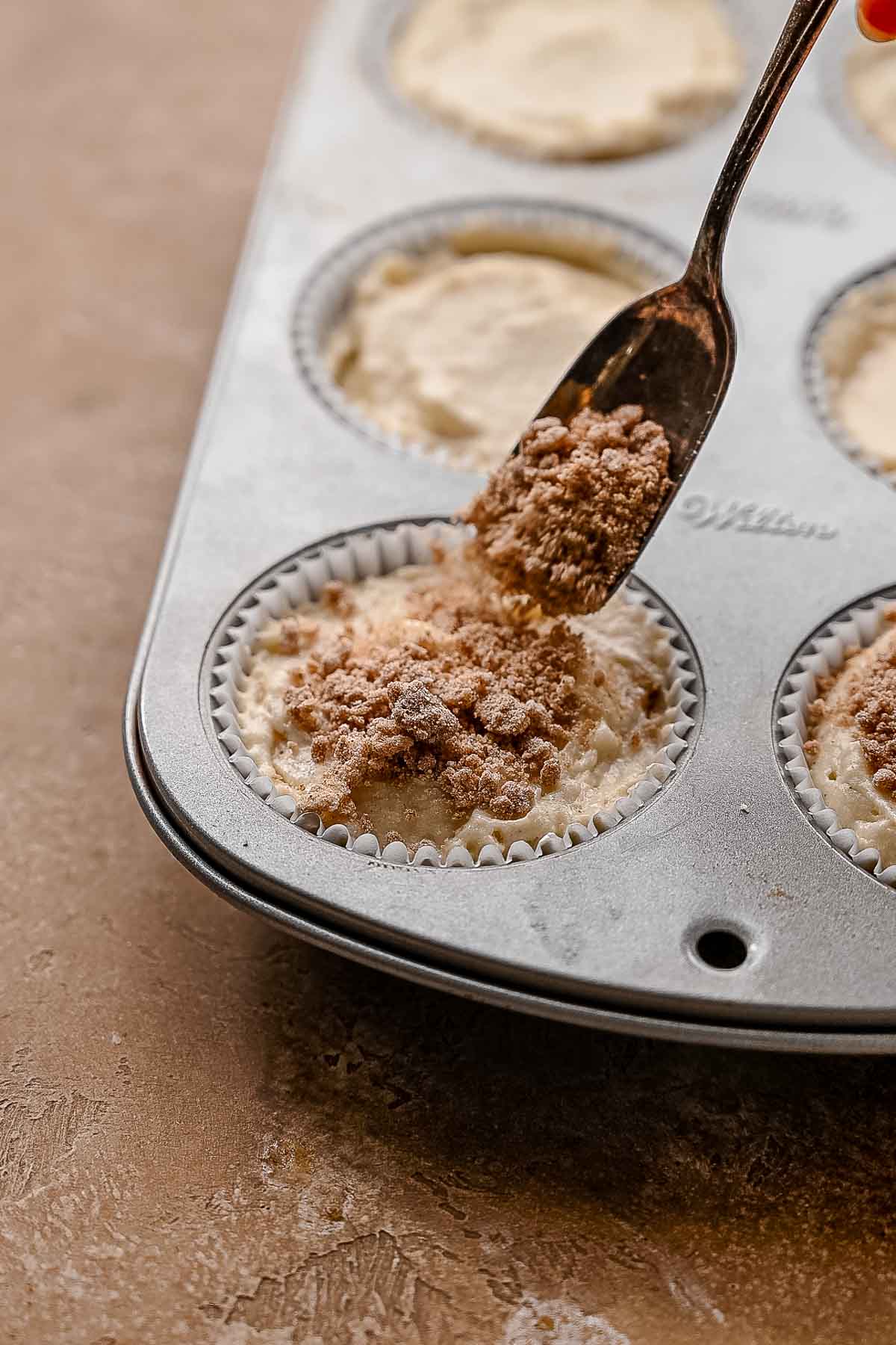Hand spooning cinnamon sugar crumble over muffin batter in a lined muffin tin before baking.