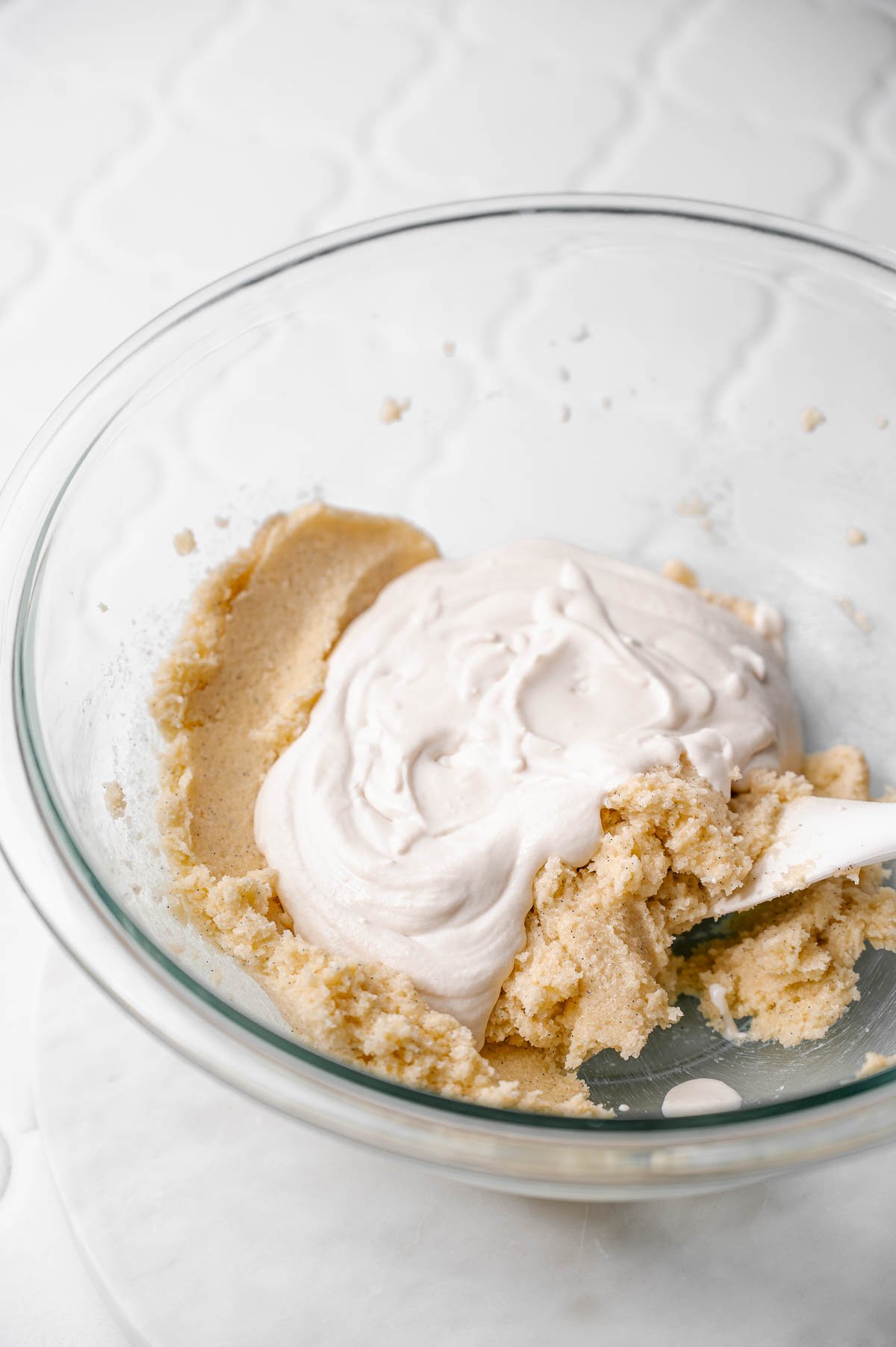 Vegan sour cream being added to creamed butter and sugar mixture in a glass mixing bowl.