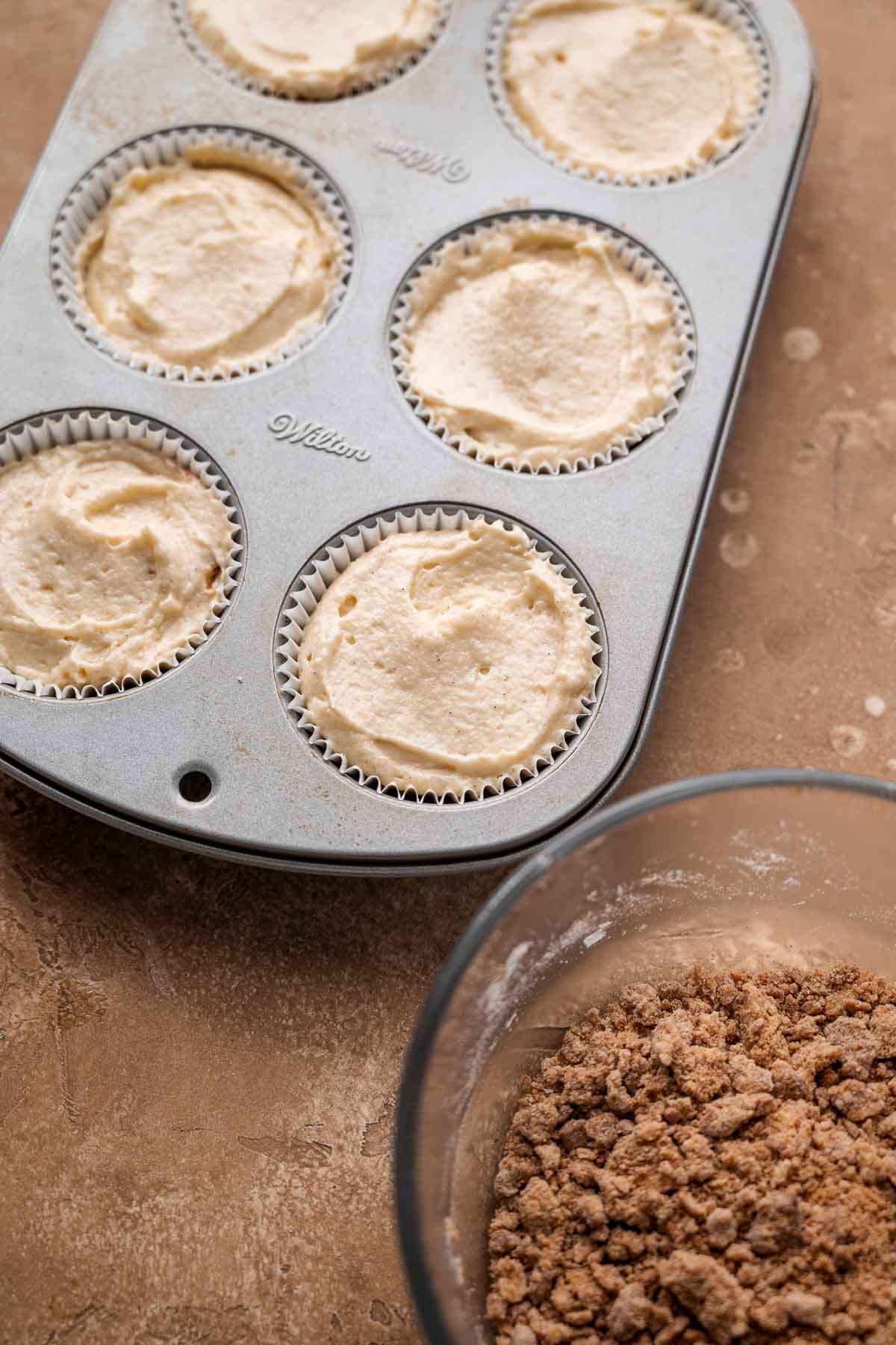 Muffin liners filled with vegan coffee cake muffin batter, ready for the crumble topping.