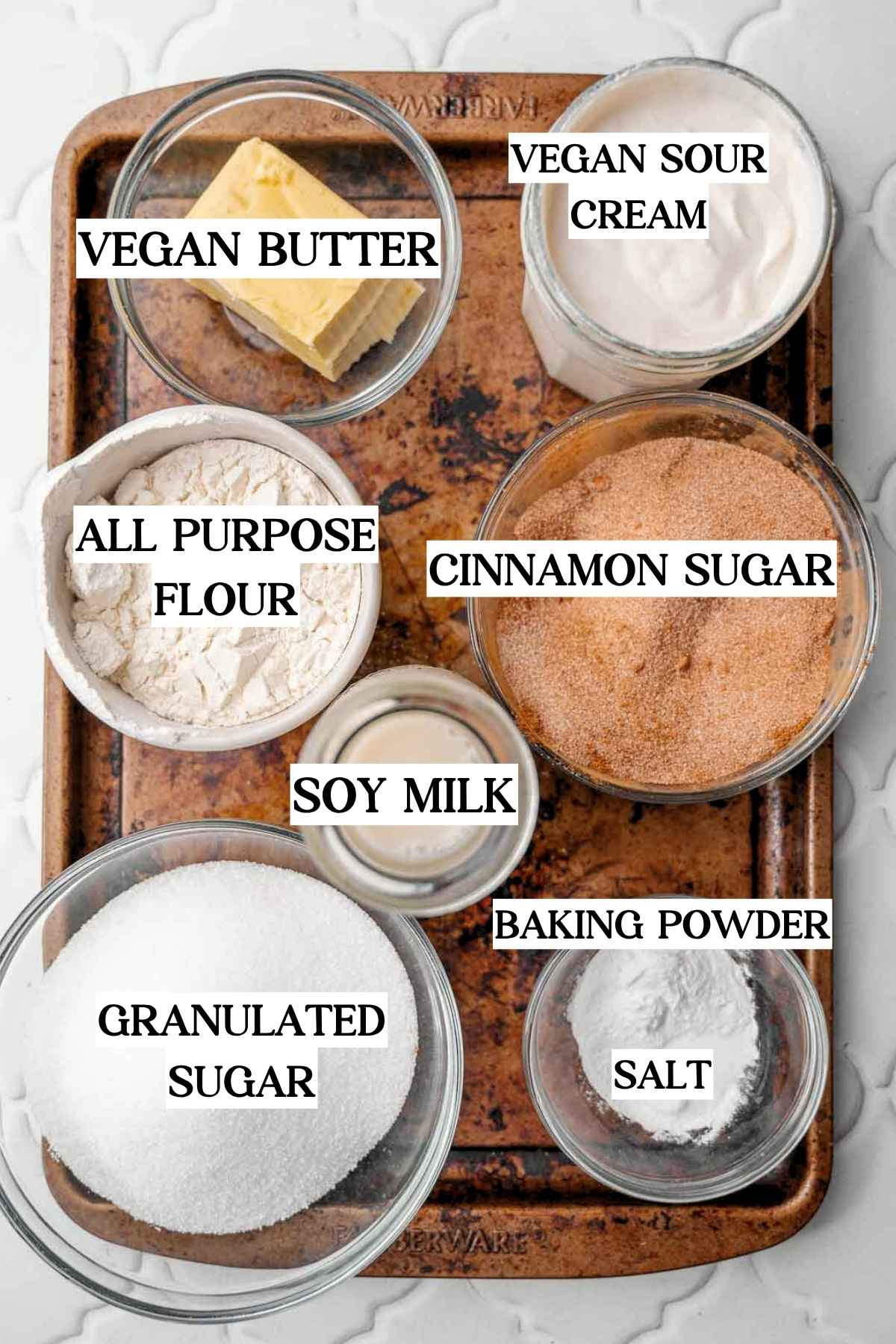 Overhead view of labeled vegan coffee cake muffin ingredients on a baking tray, including vegan butter, dairy-free sour cream, all-purpose flour, cinnamon sugar, soy milk, sugar, baking powder, and salt.
