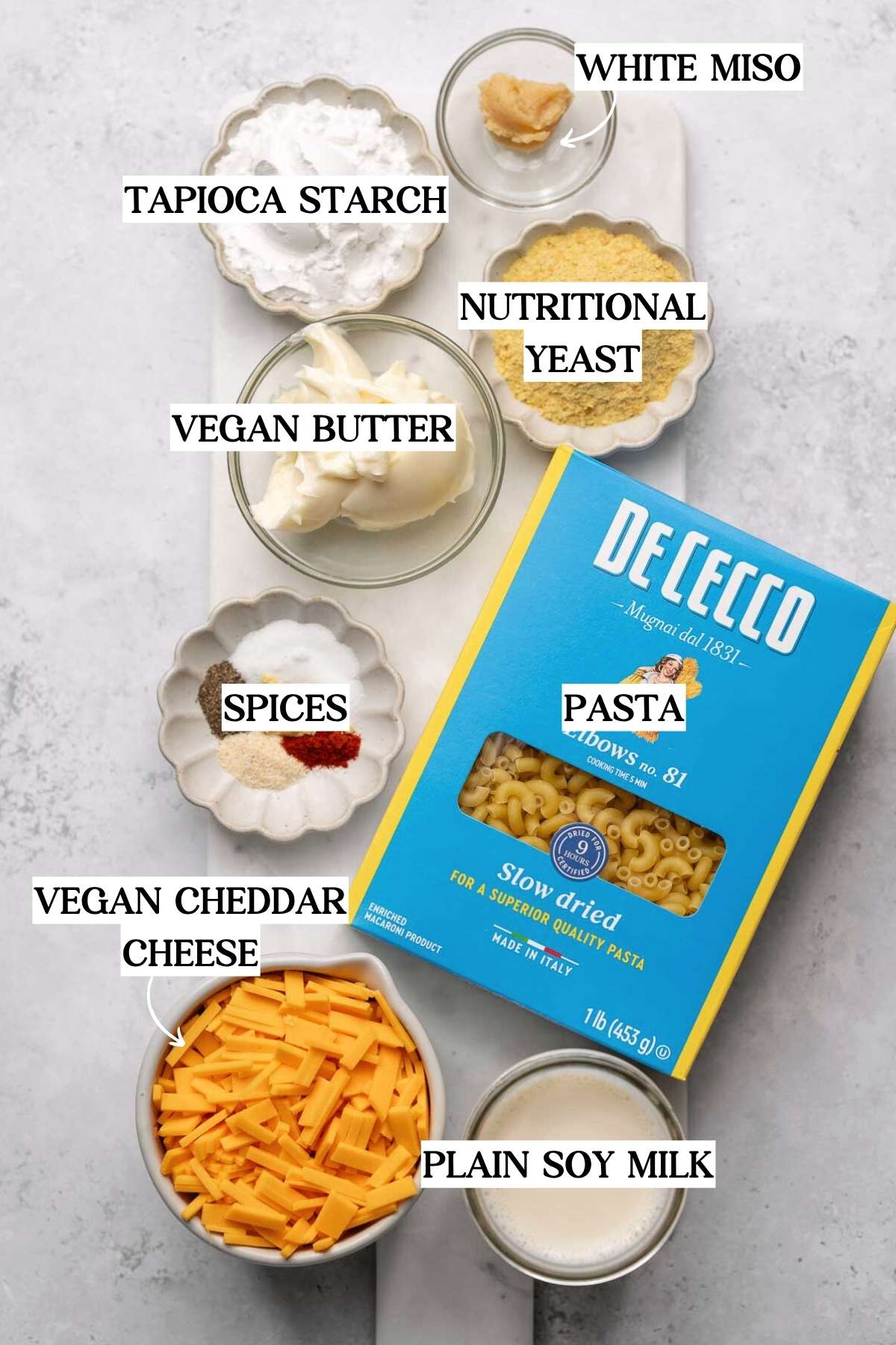 Flat lay of ingredients for vegan mac and cheese, including dry elbow pasta (De Cecco brand), plain soy milk, vegan cheddar cheese shreds, vegan butter, white miso paste, tapioca starch, nutritional yeast, and a small dish of spices including garlic powder, onion powder, paprika, and salt.