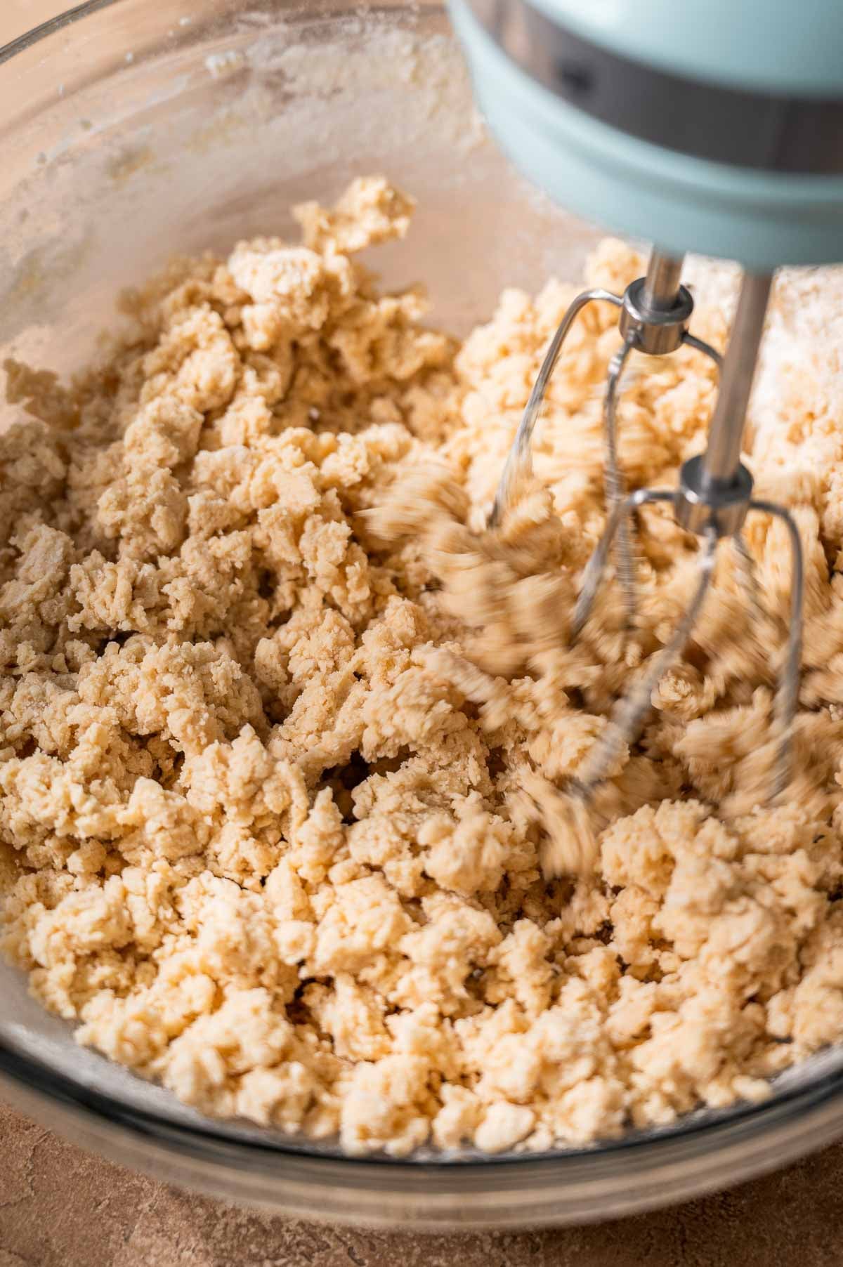 Electric mixer blending vegan cookie dough in a glass bowl until crumbly and combined.