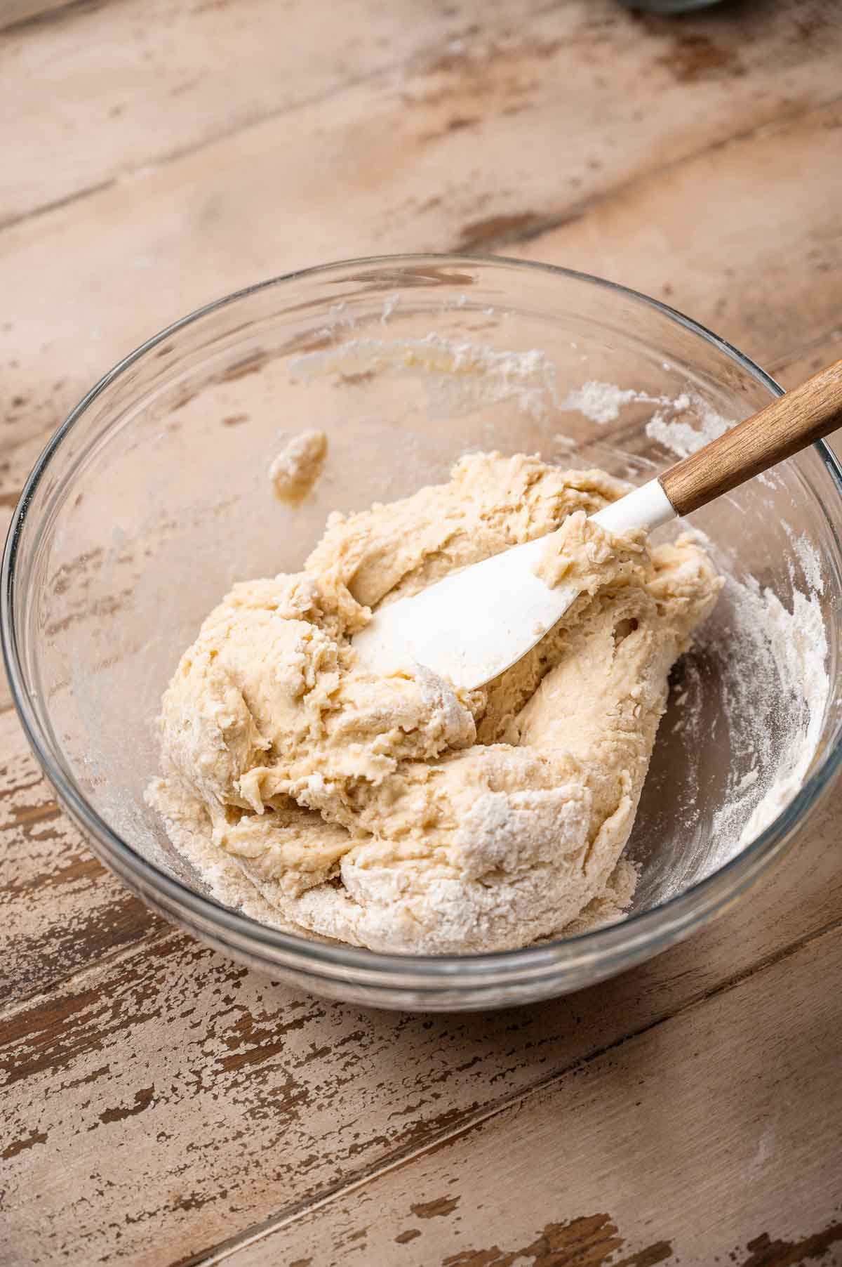 a white spatula kneading the dinner roll dough in a large glass bowl.