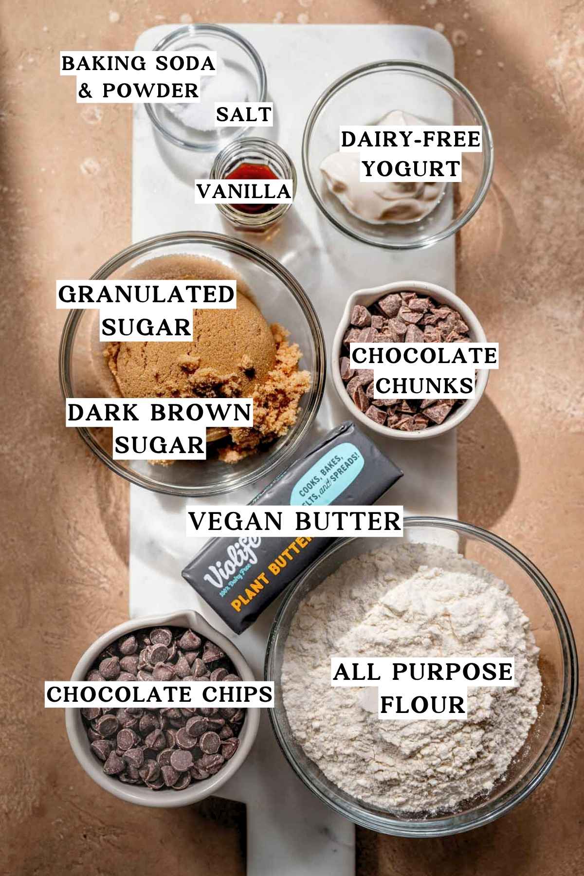 Flat lay of vegan chocolate chip cookie ingredients labeled with text overlays: vegan butter, sugars, dairy-free yogurt, vanilla, flour, baking powder and soda, salt, and chocolate chips and chunks.
