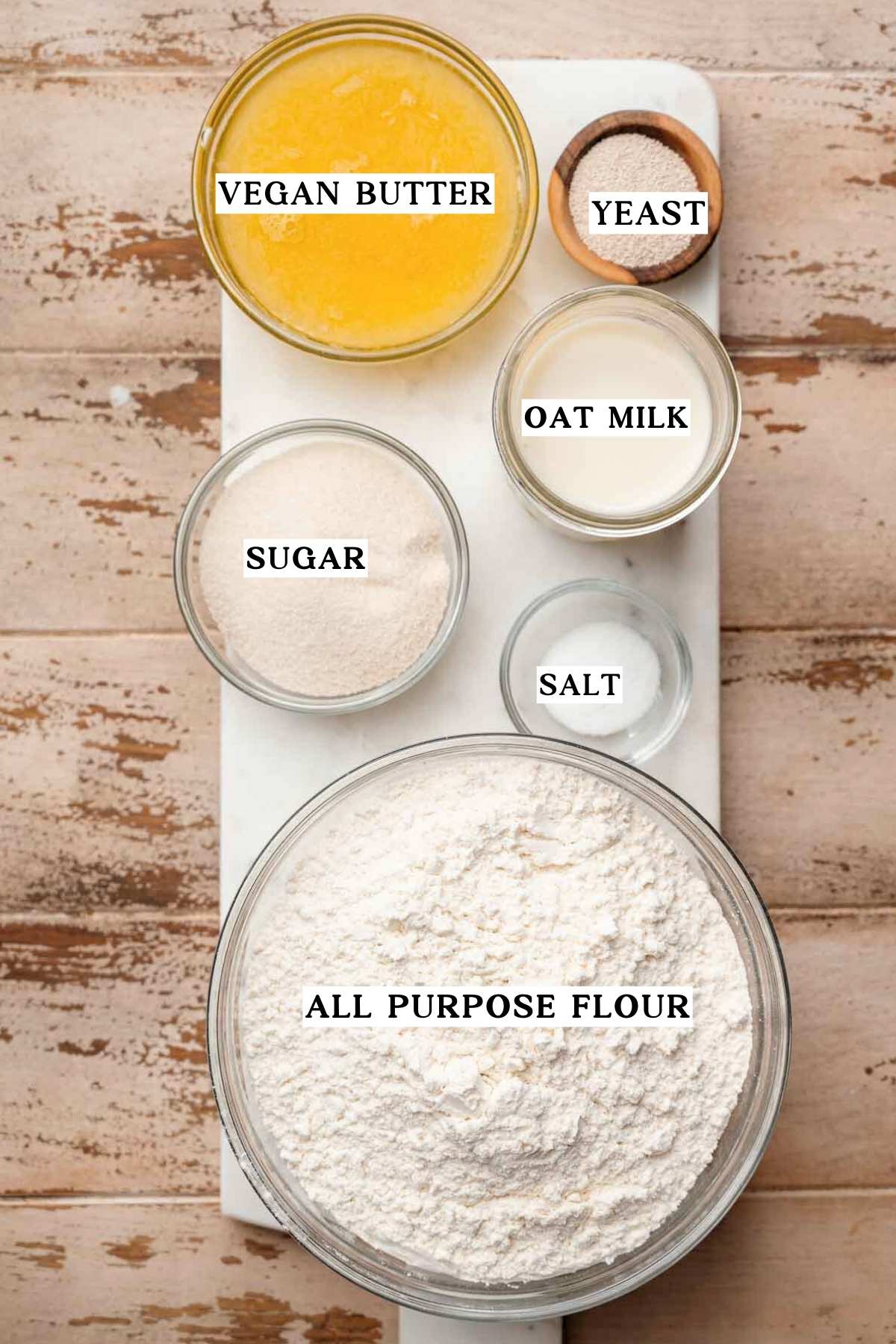 a marble cutting board with bowls of ingredients like melted vegan butter, flour, sugar and yeast.