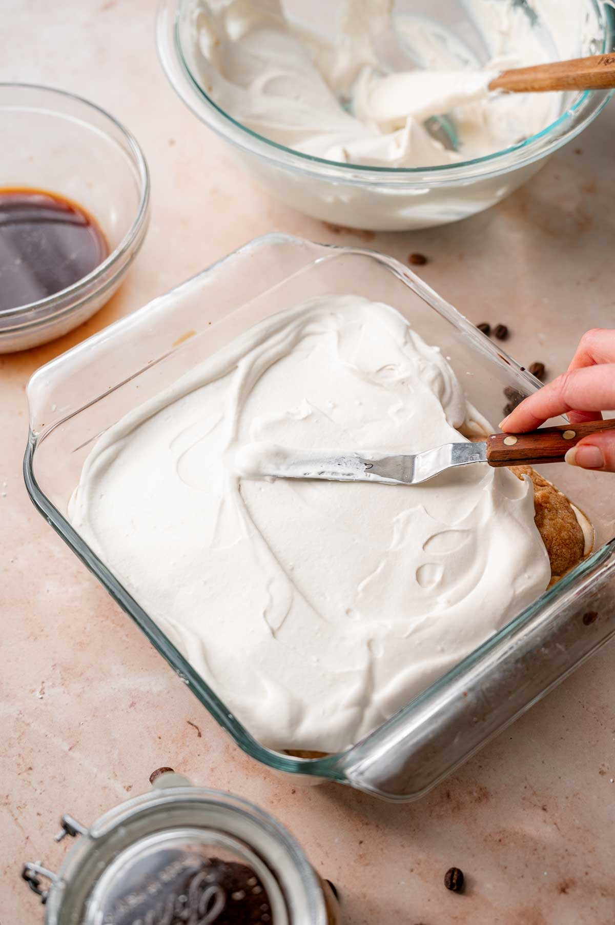 a hand using an offset spatula to smooth out the vegan mascarpone on top of the ladyfingers.