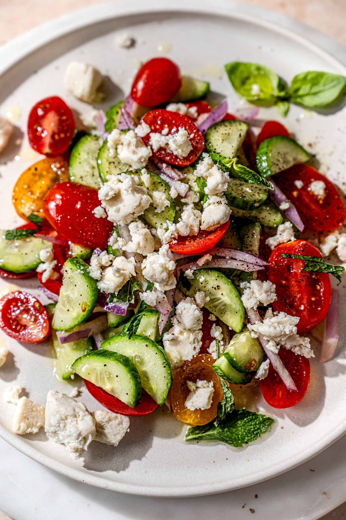 Vegan feta crumbles scattered over a fresh tomato and cucumber salad with red onion, herbs, and olive oil.